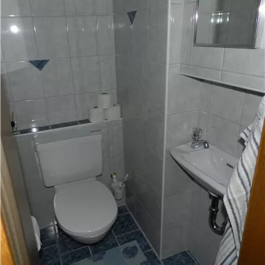 Ein kleines Badezimmer mit weißen Fliesen und blauen Akzenten. Es enthält eine Toilette, ein Waschbecken und einige Toilettenrollen.