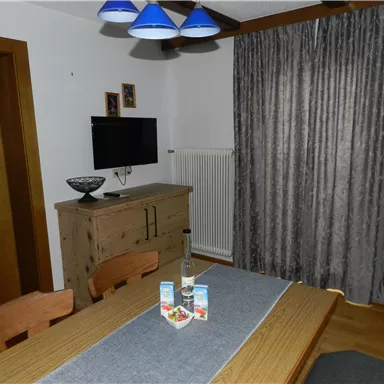 Ein gemütlicher Essbereich mit einem Holztisch und Stühlen. An der Wand hängt ein Fernseher und die Fenster sind mit Vorhängen dekoriert.