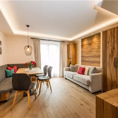 A modern living room with a stylish dining table and cozy seating. Large windows provide plenty of daylight and a warm atmosphere.