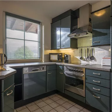 Eine moderne Küche mit dunkelblauen Schränken und Edelstahlgeräten. Durch das Fenster scheint Tageslicht herein.