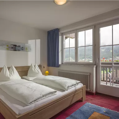 Ein gemütliches Schlafzimmer mit einem Doppelbett und zwei Kissen. Große Fenster bieten einen Blick auf die Berge und viel Tageslicht.