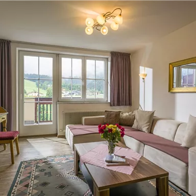 Ein gemütliches Wohnzimmer mit einem hellen Sofa und einem Tisch. Große Fenster bieten einen Blick auf die grüne Landschaft draußen.