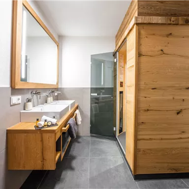 A modern bathroom with wooden elements and a large mirror. The shower is separated by a glass wall and the floor is dark tiled.