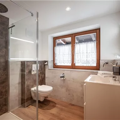 Ein modernes Badezimmer mit einer Glasdusche und einem weißen Waschbecken. Großen Fenstern lassen viel Licht herein.