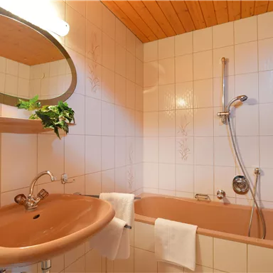 Ein Badezimmer mit einer Badewanne und einem Waschbecken. Die Wände sind hell gefliest und ein runder Spiegel hängt an der Wand.