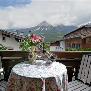 Ein eleganter Balkon mit einem runden Tisch und Stühlen. Im Hintergrund sind Berge und charmante Holzhäuser zu sehen.
