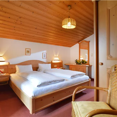 A cozy bedroom with wooden walls and sloped ceilings. Two beds, one chair, and soft lighting create an inviting atmosphere.