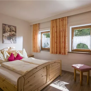 Ein gemütliches Schlafzimmer mit einem großen Bett und bunten Kissen. Helle Vorhänge lassen viel Licht herein und eine kleine Blumenvasen sorgt für eine freundliche Atmosphäre.