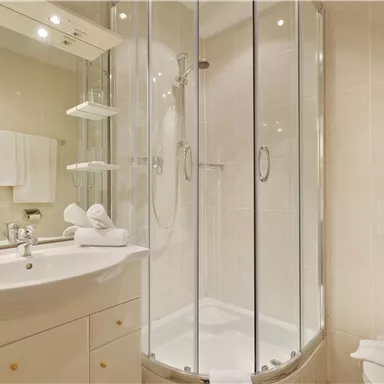 Ein modernes Badezimmer mit einer Glaskabine für die Dusche. Es gibt ein Waschbecken, Handtücher und helle Fliesen.