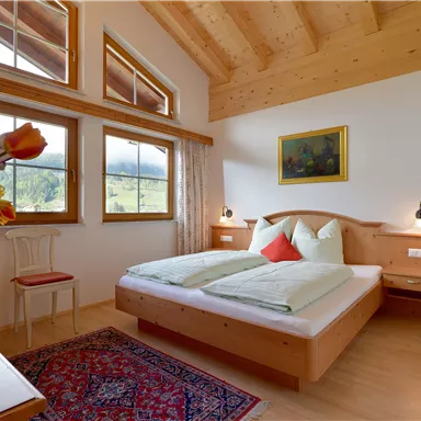 Ein gemütliches Schlafzimmer mit Holzdecke und großen Fenstern. Die Einrichtung ist hell und einladend, mit einem Farbtupfer durch Kissen und Blumen.