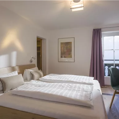A bright and modern bedroom with two single beds. There is a desk, a television, and a window with curtains.