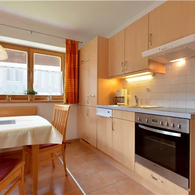 A modern kitchen with wooden cabinets and a dining table. The windows are decorated with orange curtains and the room is well-lit.