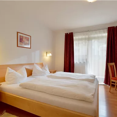 A cozy bedroom with two beds, a table, and chairs. Large windows provide plenty of daylight.