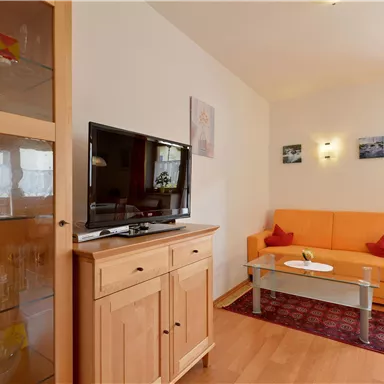 A cozy living room with an orange couch and a television. There is a glass display cabinet and some pictures on the wall.