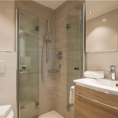 A modern bathroom with a shower and a sink. The walls are tiled in light colors, and there are towels made of white cotton.