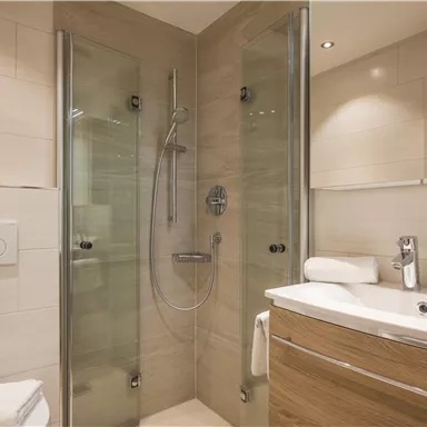 A modern bathroom with a shower and a sink. The walls are tiled in light colors, and there are towels made of white cotton.