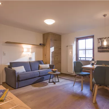 A modern living room with a blue sofa and a dining table. Large windows with a view outside and wooden elements create a cozy atmosphere.