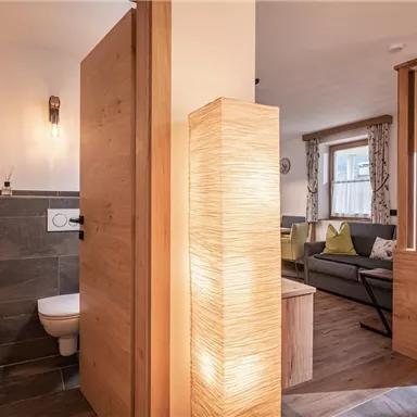 A modern bathroom with a toilet and wooden details. In the background, a bright living space with cozy furniture is visible.