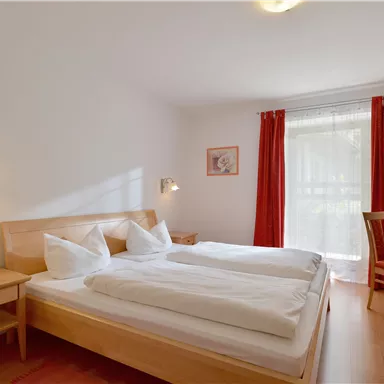A cozy bedroom with two single beds and light wooden furniture. Large windows with red curtains let in plenty of daylight.