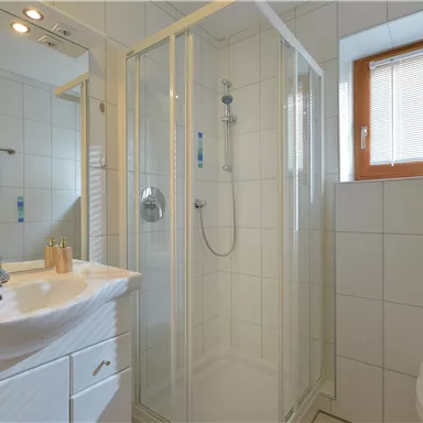 A modern bathroom with a shower cabin and a sink. The walls are tiled white and a window provides natural light.