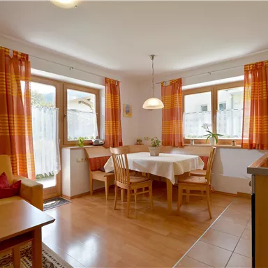 A cozy kitchen with a dining area and wooden chairs. Bright windows with orange curtains let in plenty of light.