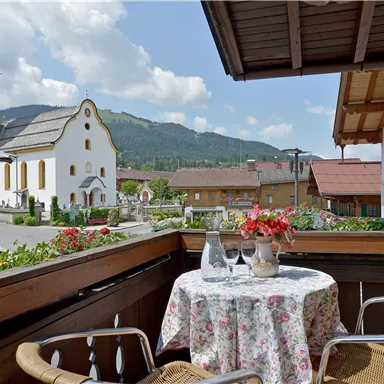 Ein Balkon mit einem Tisch und Stühlen, geschmückt mit Blumen. Im Hintergrund ist eine Kirche und die umliegenden Berge zu sehen.