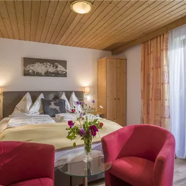 Ein gemütliches Schlafzimmer mit einem Doppelbett und rot gefütterten Sesseln. Dazu eine Blumenvase und ein Fenster mit Vorhängen, das Tageslicht hereinlässt.