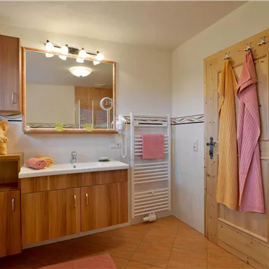 A modern bathroom with wooden cabinets and a large mirror light. There is a shower, towels in various colors, and a warm atmosphere.