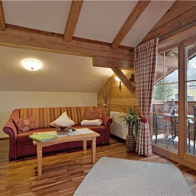 A cozy living room with wooden beams and a comfortable couch. The room has large windows overlooking a balcony and the surrounding nature.