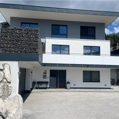 A modern building with multiple windows and a balcony. The facade is in white and gray, surrounded by a parking lot and a stone wall.