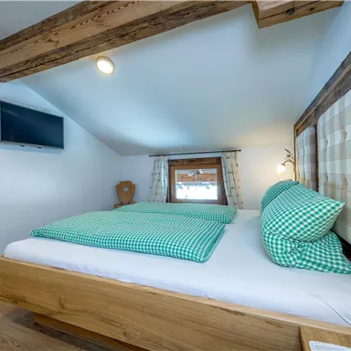 A cozy bedroom with a wooden frame bed and checked pillows. A television hangs on the wall and the room has bright, inviting colors.