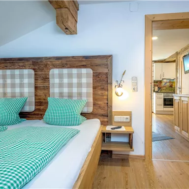 A cozy bedroom with two beds and checkered headboards. The room has wood details and a view of a kitchen through the open door.