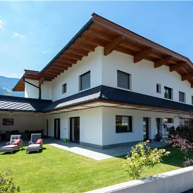 Ein modernes Haus mit einem großen Garten und einer Terrasse. Im Hintergrund sind Berge und ein klarer Himmel zu sehen.