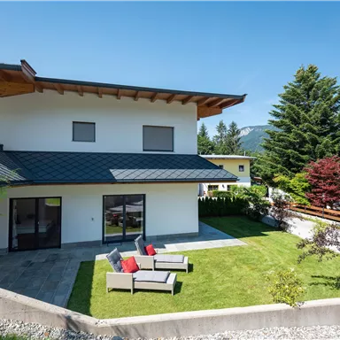 Ein modernes Haus mit einer schönen Terrasse und einem gepflegten Garten. Im Hintergrund sind Berge und Bäume zu sehen.