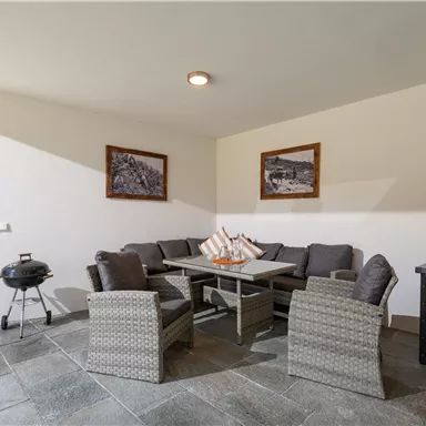 Eine gemütliche Terrasse mit modernen Möbeln und einem Tisch. Im Hintergrund sind einige dekorative Bilder an der Wand zu sehen.