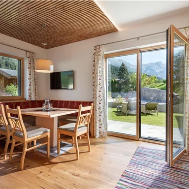 Ein gemütliches Wohnzimmer mit Holzdecke, einem Esstisch und Stühlen. Durch die offenen Fenster blickt man auf einen Garten mit Bergblick.