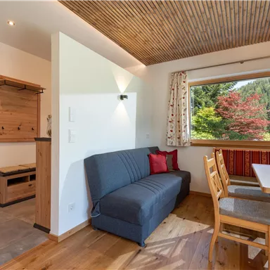 Ein gemütlicher Wohnraum mit Holzfußboden und einer modernen Sitzecke. Große Fenster bieten einen Blick auf die grüne Natur draußen.