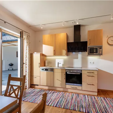 Eine moderne Küche mit Holzschränken und einer offenen Verbindung zum Essbereich. Der Raum ist hell erleuchtet und hat große Fenster, die den Blick ins Freie freigeben.