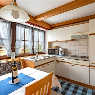 Eine helle Küche mit Holzverkleidung und großen Fenstern. Auf dem Tisch steht eine Flasche Wein und eine Tischdecke in Blau.