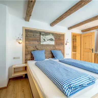 A cozy bedroom with a double bed and blue and white checked blankets. The walls are made of wood, and there are large windows with plenty of natural light.