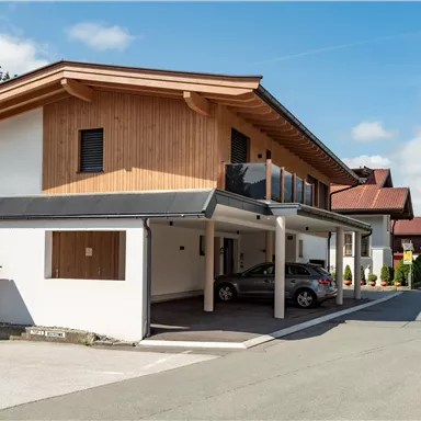 Ein modernes Gebäude mit Holzverkleidung und einem überdachten Parkplatz. Die Straße ist ruhig und von anderen Häusern gesäumt.