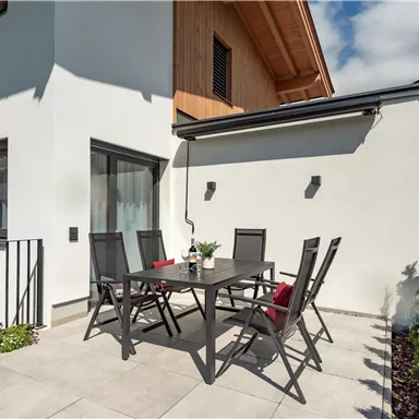 Ein eleganter Außenbereich mit einem Tisch und Stühlen. Die Terrasse ist hell und einladend, umgeben von grünen Pflanzen.