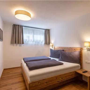 Ein modernes Schlafzimmer mit einem Doppelbett und Holzdetails. Der Raum ist hell und freundlich mit einem Fenster und einem Fernseher.