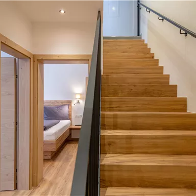 Ein moderner Flur mit einer Holztreppe und einem Geländer. Im Hintergrund ist ein Schlafzimmer mit gemütlicher Beleuchtung zu sehen.