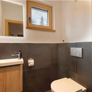 Ein modernes Badezimmer mit einer eleganten Toilette und einem kleinen Waschbecken. Große Fenster sorgen für viel Licht und eine freundliche Atmosphäre.