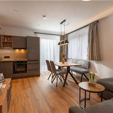 Ein modernes Wohnzimmer mit einer offenen Küchenzeile und einem Essbereich. Helle Farben und große Fenster sorgen für eine freundliche Atmosphäre.