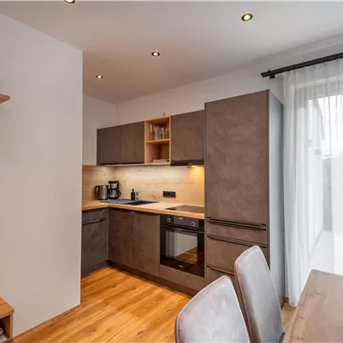 Eine moderne Küche mit Holzfußboden und eleganten Möbeln. Sie ist gut beleuchtet und verfügt über Fenster mit Vorhängen sowie eine Essgelegenheit.