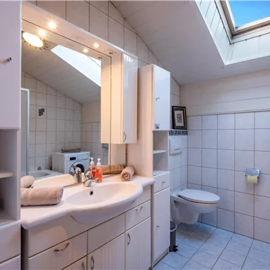 A bright bathroom with a sink, a large mirror, and a window. The walls are tiled, and there are towels as well as toiletries.