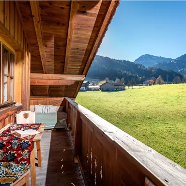 A cozy balcony with wooden cladding and a view of a green meadow and mountains in the background. The sky is clear and sunny.