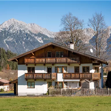 A beautifully situated house in the mountains with a large garden. Majestic mountains and a blue sky are visible in the background.